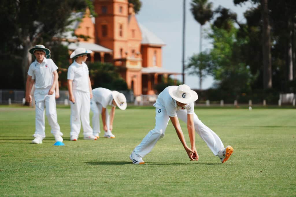 Student cricketers in white uniform on a sports field during training.