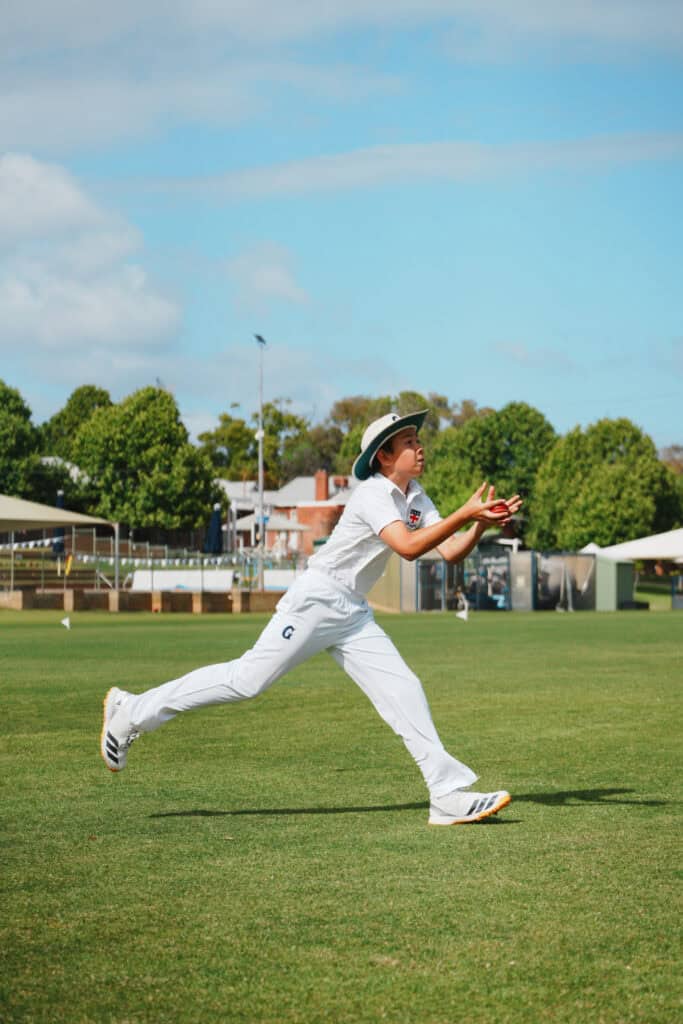 Cricketer in white uniform leaping to catch on a sunny sports field.