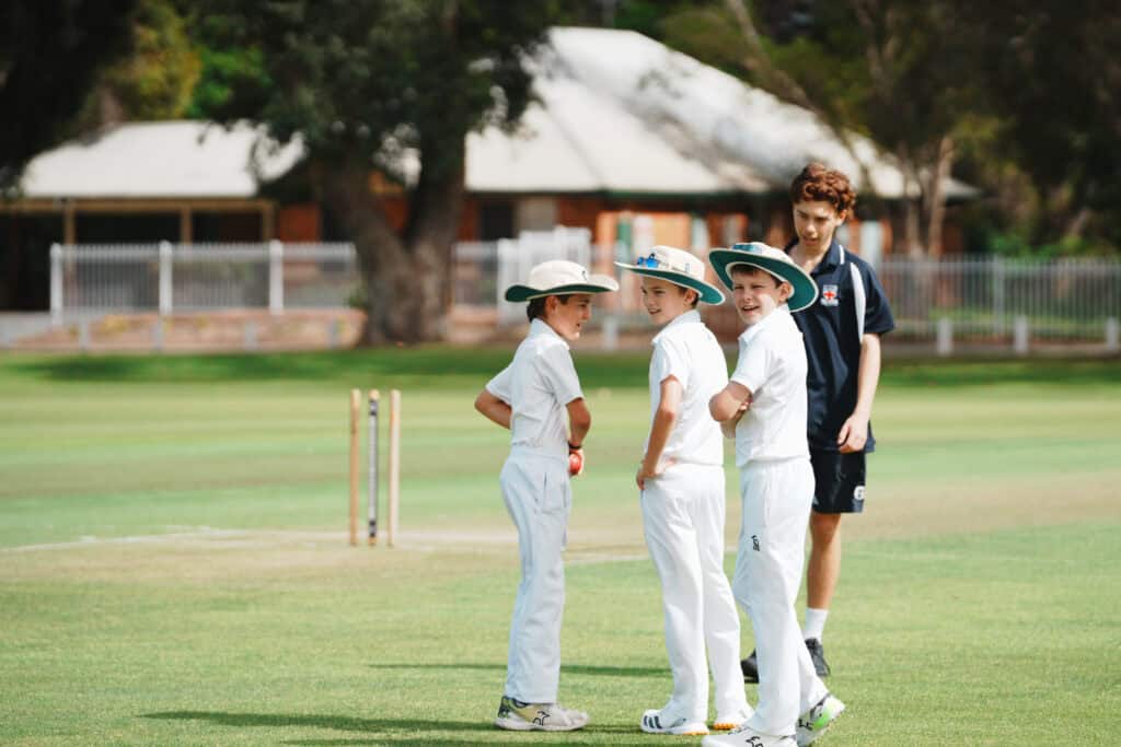 Group of young cricketers in white uniforms listening to coach on a sunny field.