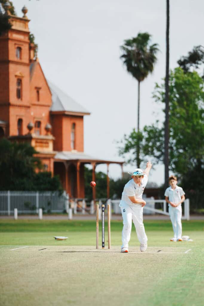 Cricket bowler in white uniform delivering a ball on a green field with a historic red-brick wall in.