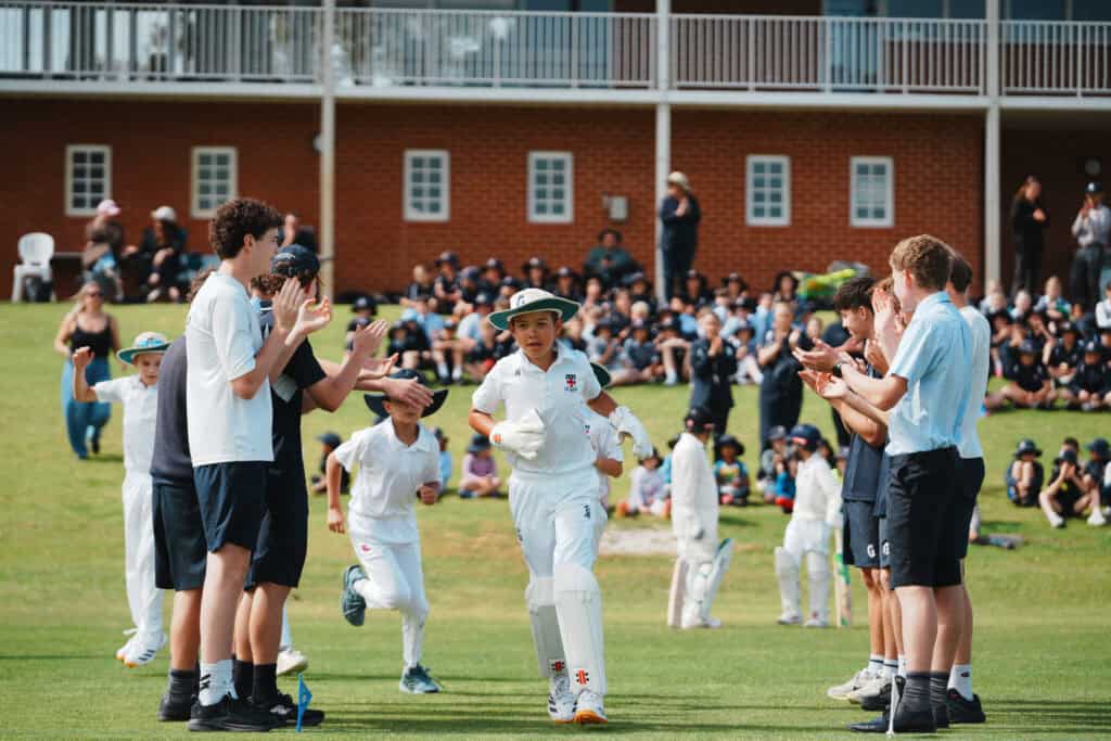 Group of students in cricket whites applauding on a school sports field.